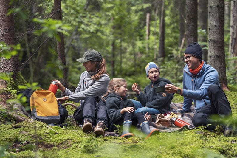 Familj på en utflykt i skogen där barnen bär Husqvarna Xplorer Kids luvtröjor. De sitter på en mossig mark och delar en måltid, med skogen som en naturskön bakgrund.