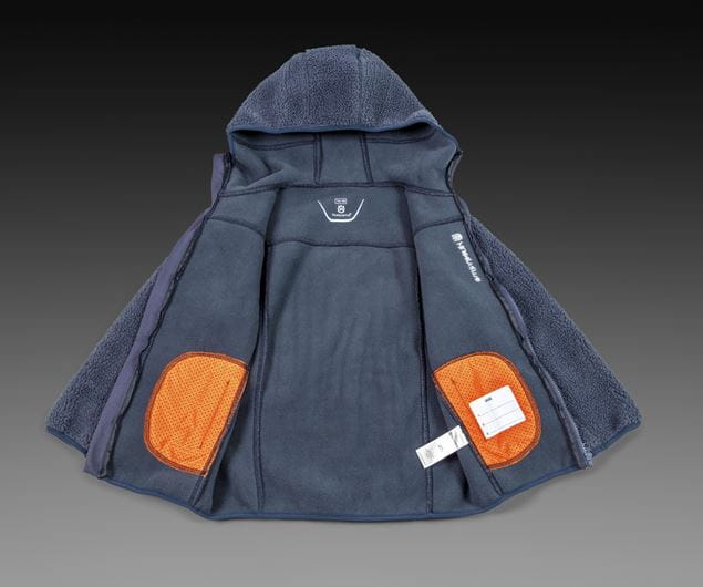 En blå Husqvarna Xplorer Kids luvtröja läggs platt på en mörk bakgrund, visar tröjans insida med orangea lappar på varje sida, en logotyp upptill och en vit etikett längst ner